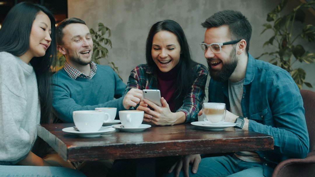 Four friends laughing together at a cafe table while reading a slang message on a smartphone